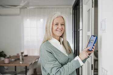 Glückliche reife Frau, die den Thermostat mit ihrem Smartphone zu Hause einstellt - EBBF07651