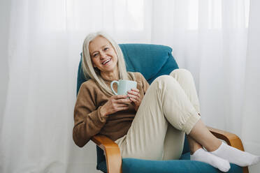 Happy mature woman with coffee cup sitting on armchair at home - EBBF07654