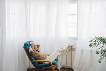 Woman relaxing on armchair in front of window at home - EBBF07681