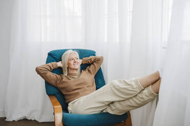 Smiling mature woman with hands behind head relaxing on armchair - EBBF07682