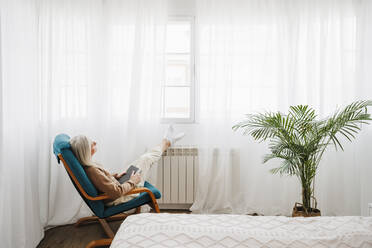 Thoughtful woman with book sitting on armchair at home - EBBF07684