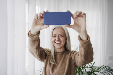 Glückliche reife Frau, die zu Hause mit ihrem Smartphone fotografiert - EBBF07707