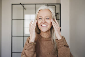 Glückliche reife Frau, die zu Hause Feuchtigkeitscreme auf ihr Gesicht aufträgt - EBBF07713