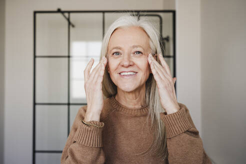 Happy mature woman applying moisturizer on face at home - EBBF07713