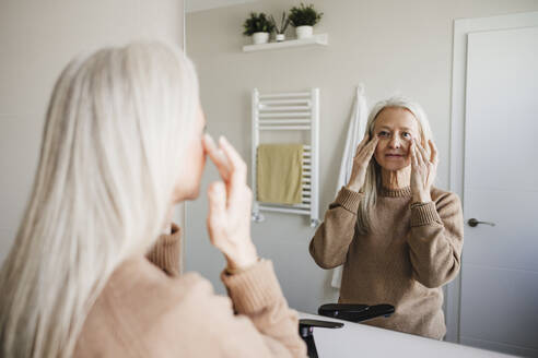 Reife Frau schaut in den Spiegel und trägt Feuchtigkeitscreme auf das Gesicht auf - EBBF07714