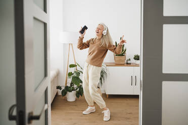 Cheerful mature woman wearing wireless headphones dancing at home - EBBF07721