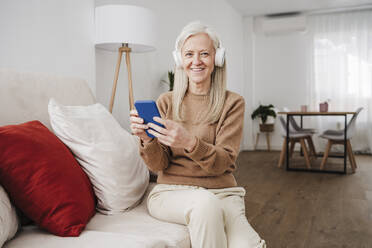 Glückliche reife Frau mit Smartphone auf dem Sofa sitzend - EBBF07725