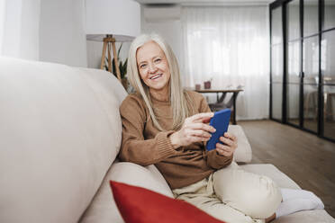 Glückliche reife Frau sitzt mit Handy zu Hause - EBBF07728