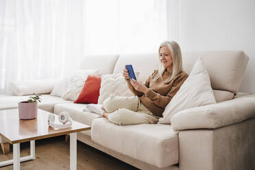 Smiling mature woman using smart phone sitting on sofa at home - EBBF07729