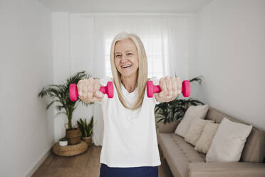 Happy woman practicing exercise with dumbbells in living room - EBBF07735