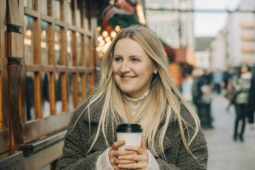 Glückliche blonde Frau mit Kaffeetasse auf dem Fußweg auf dem Weihnachtsmarkt - VSNF00317