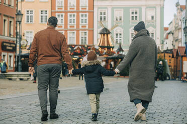 Mother and father holding hands with son and walking on footpath at Christmas market - VSNF00320
