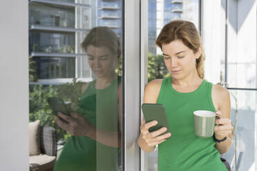 Woman holding cup using smart phone leaning on window - SVKF01049