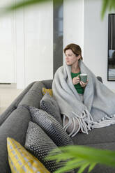 Nachdenkliche, in eine Decke eingewickelte Frau sitzt zu Hause auf dem Sofa - SVKF01061
