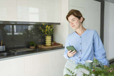 Thoughtful woman standing with smart phone in kitchen - SVKF01062