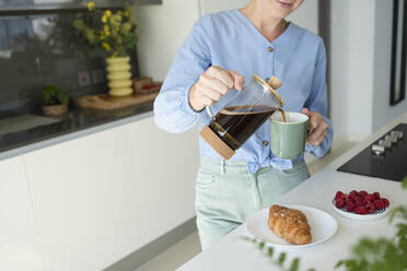 Frau gießt Kaffee in Tasse mit Croissant auf dem Tresen zu Hause - SVKF01063
