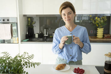 Frau hält Kaffeetasse mit Croissant auf dem Tresen in der Küche - SVKF01066