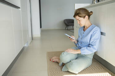 Woman using smart phone sitting by counter in kitchen - SVKF01068