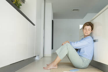 Frau mit geschlossenen Augen sitzt auf dem Boden neben dem Smartphone in der Küche - SVKF01069