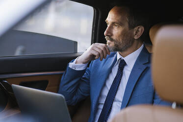 Thoughtful businessman sitting with laptop in backseat of car - OIPF02753