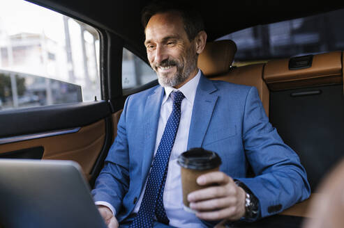 Glücklicher reifer Geschäftsmann mit Laptop und Einweg-Kaffeebecher im Auto - OIPF02756