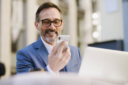 Glücklicher reifer Geschäftsmann mit Blick auf sein Smartphone im Café - OIPF02772