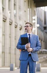Thoughtful mature businessman with clipboard on footpath in front of building - OIPF02783