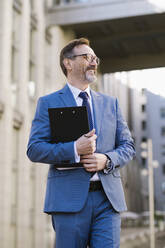 Happy mature businessman with clipboard standing in front of building - OIPF02784