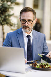 Reifer Geschäftsmann bei der Arbeit und beim Mittagessen vor einem Laptop in einem Cafe - OIPF02788