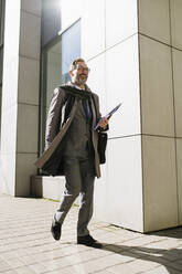 Happy mature businessman with clipboard walking on footpath at sunny day - OIPF02799