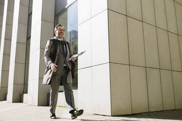 Smiling businessman with clipboard walking on footpath outside office building - OIPF02801