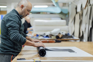 Upholstery worker rolling fabric by table in workshop - PGF01442