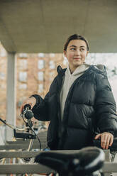 Smiling teenage girl in black jacket standing at bicycle parking station - VSNF00325