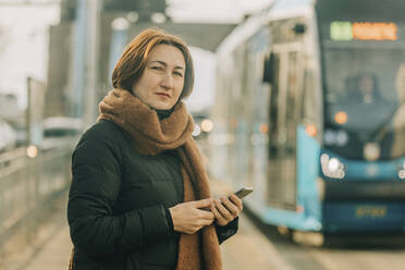 Lächelnde reife Frau steht mit Smartphone an der Straßenbahnhaltestelle - VSNF00337