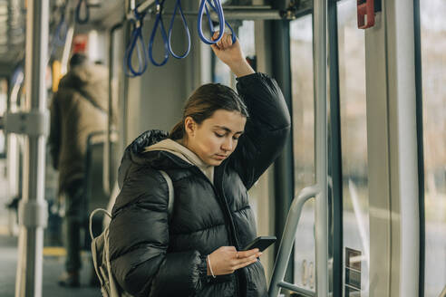 Teenage girl using smart phone standing in tram - VSNF00342