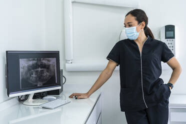 Arzt mit chirurgischer Maske betrachtet ein Röntgenbild auf einem Computer in einer Klinik - DLTSF03524