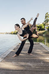 Man lifting woman practicing ballet dance on wooden pier - JRVF03286