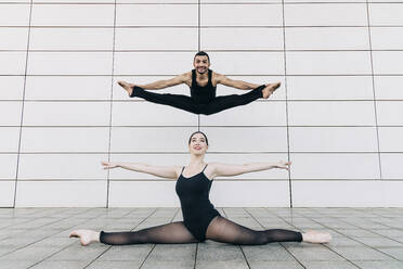 Happy man and young woman doing splits in front of tiled wall - JRVF03296