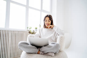 Happy woman using laptop sitting cross-legged at home - MDOF00493