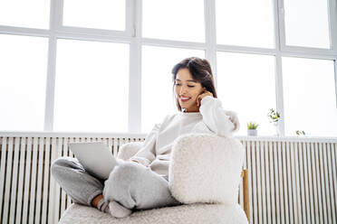 Happy young woman using laptop sitting by radiator at home - MDOF00495