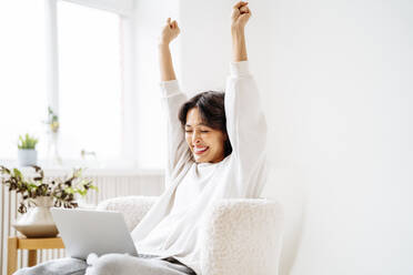 Excited young woman with arms raised sitting on chair at home - MDOF00509