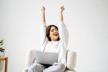Excited woman with arms raised sitting on chair at home - MDOF00510