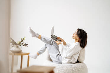 Frau, die einen Laptop benutzt, sitzt auf einem Stuhl an der Wand zu Hause - MDOF00514
