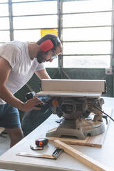 Zimmermann mit Gehörschutz, der in einer Werkstatt ein Brett mit einer Kreissäge schneidet - JAQF01203