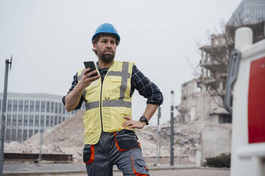Arbeiter hält Smartphone mit Hand auf der Hüfte vor einer Baustelle - JOSEF15748