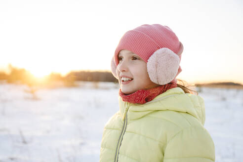 Happy girl wearing knit hat and ear muff in winter forest - EYAF02519