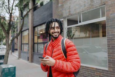 Smiling man using smart phone standing in front of building - DSIF00658