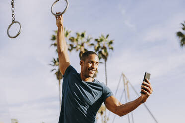 Glücklicher Mann, der mit Gymnastikringen trainiert und ein Selfie mit seinem Smartphone macht - OIPF02812