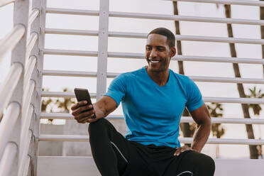 Happy man using smart phone sitting in front of railing - OIPF02862