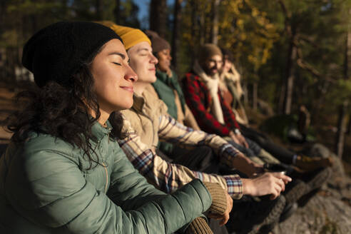 Seitenansicht einer lächelnden Frau mit geschlossenen Augen, die das Sonnenlicht genießt, während sie bei Freunden im Wald sitzt - MASF34186